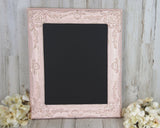 An ornate wood and plaster frame painted in distressed pink and gold layers surrounding a clean black chalkboard vinyl surface.