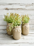 Hand-painted light brown country mason jars filled with artificial white floral sprays acting as centerpieces on a wooden wedding reception table.